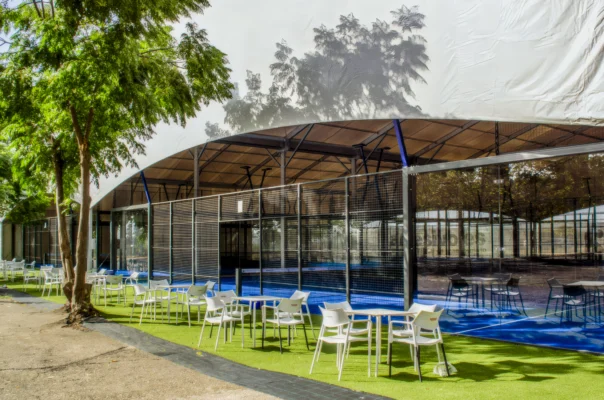 Terraza exterior con mesas y sillas junto a una pista deportiva acristalada rodeada de árboles.